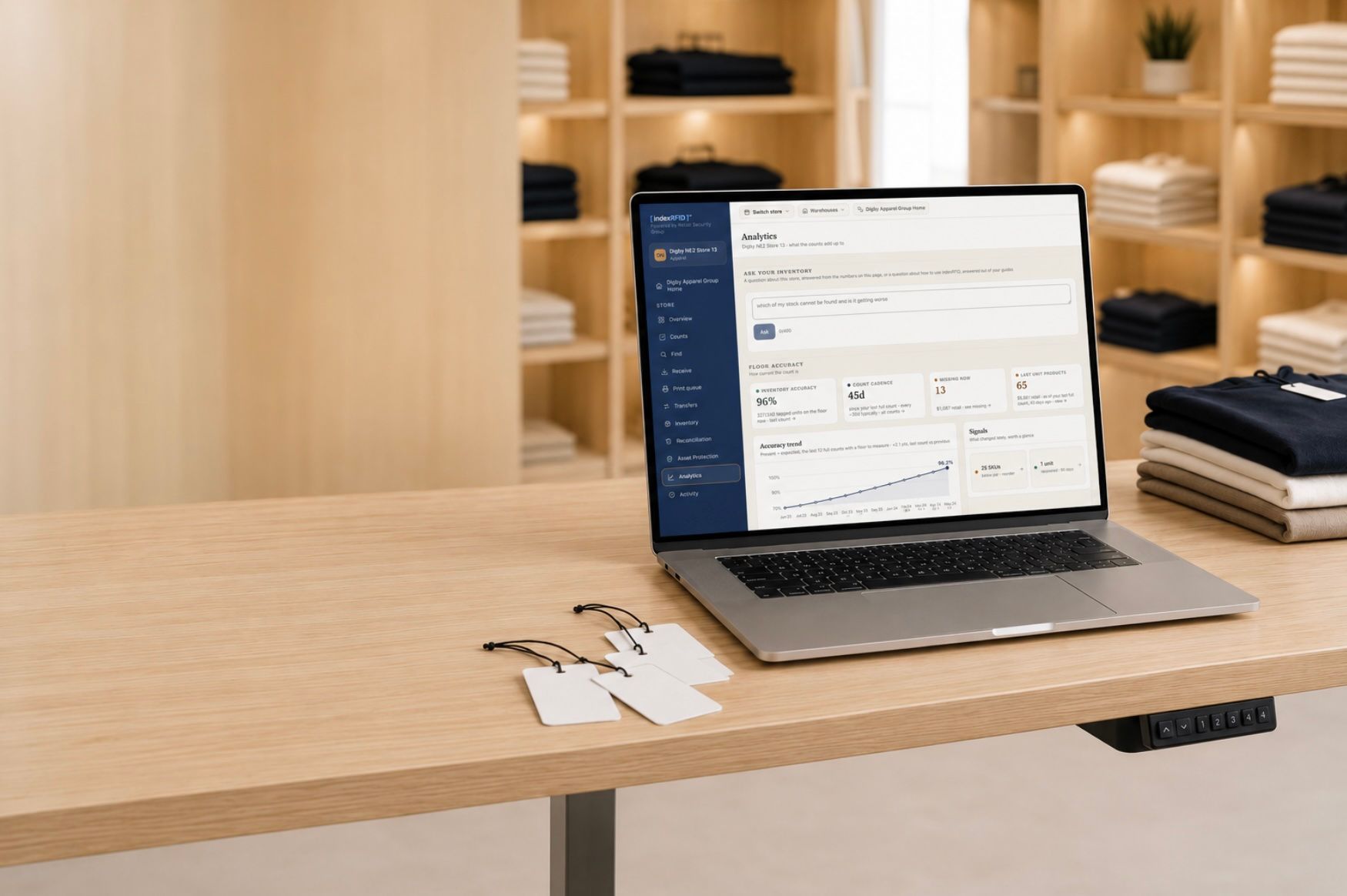 A laptop on a wood table in a clothing store showing the indexRFID Analytics screen with the Ask your inventory box, RFID hangtags and folded knitwear beside it